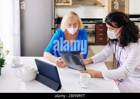 Junge Ärztin in medizinischer Maske besucht Seniorpatientin zu Hause und zeigt ihre Lungen Röntgen Stockfoto
