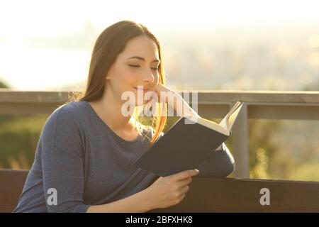 Entspannte Frau, die bei Sonnenuntergang ein Hardcover liest und auf einer Parkbank sitzt Stockfoto