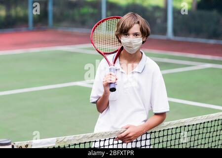 Junge in Gesicht Maske Tennis spielen auf dem Außenplatz während Coronavirus Ausbruch. Teenager mit Tennisschläger und Ball im Sportclub. Aktive Übung für Stockfoto