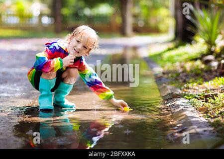 Kind spielt im Regen im Herbstpark. Kind springt in schlammigen Pfütze an regnerischen Herbsttag. Kleiner Junge in Regenstiefeln und roter Jacke im Freien in schweren sho Stockfoto