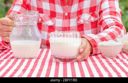 Frau in einem karierten roten Hemd hält ein Glas Milch. Auf dem Tisch ist eine rote Tischdecke. Auf der Tischdecke befindet sich ein Krug mit Milch und saurer Sahne. Beschaffung Stockfoto