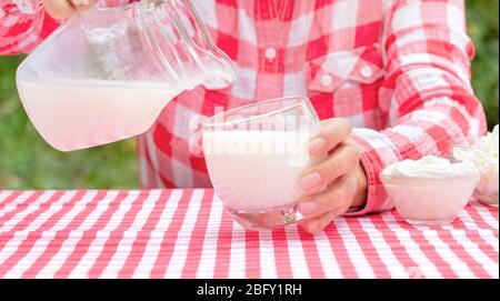 Frau gießt Milch aus einem Krug in ein Glas an einem Tisch mit einer roten Tischdecke. Auf dem Tisch sind Milchprodukte. Natürlicher grüner Hintergrund Stockfoto