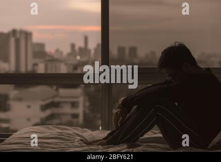 Der Mann, der auf dem Bett sitzt und vom Schlafzimmer aus die Stadt betrachtet. Bleiben Sie zu Hause, Depression und Einsamkeit Konzept. Stockfoto