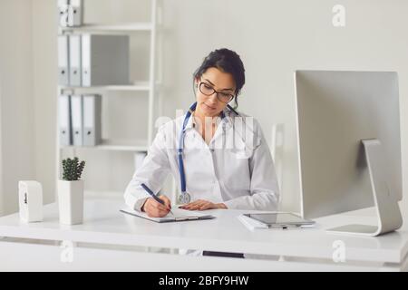 Selbstbewusste Frau Ärztin Kinderärztin schreibt in einer Zwischenablage, die an einem Tisch in einem weißen Büro des Krankenhauses sitzt Stockfoto