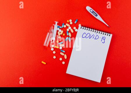 Notizblock, eine Vielzahl von Medikamenten, Thermometer, Spritze und Ampullen mit Medizin auf rotem Hintergrund. Blau auf einem Notizbuch COVID-19 geschrieben. Stockfoto