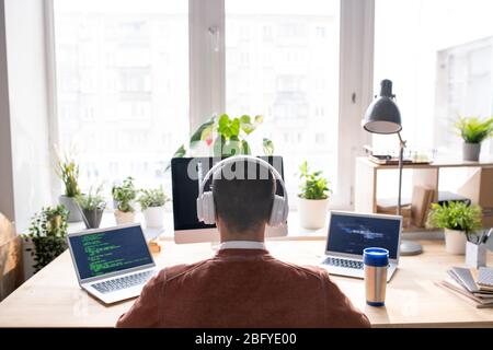 Rückansicht des Computerprogrammierers in drahtlosen Kopfhörern, die am Tisch sitzen und mit drei Computern am Code arbeiten Stockfoto