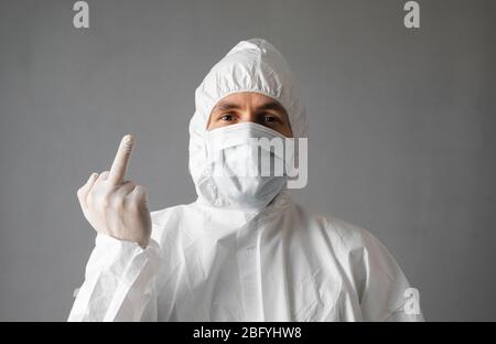 Der Arzt in weißem Schutzanzug, medizinischer Maske und Gummihandschuhen zeigt dem Virus Mittelfinger. Hilfe für Menschen während einer Coronavirus-Pandemie Stockfoto