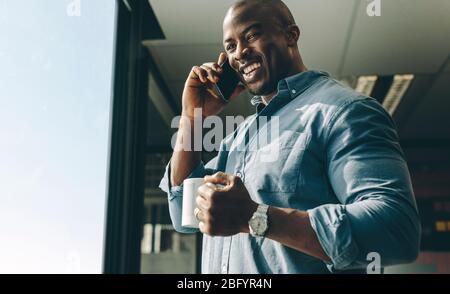 Glücklicher junger Geschäftsmann mit Kaffee in der Hand, der im Handy-Büro spricht. Geschäftsmann lächelt, während er telefoniert. Stockfoto