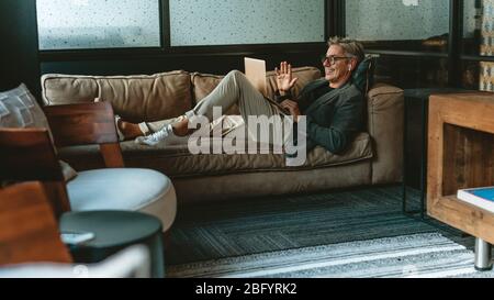 Geschäftsmann, der einen Videoanruf mit einem Laptop-Computer macht. Männlicher Profi, der im Büro auf dem Sofa liegt und einen Videoanruf macht. Stockfoto