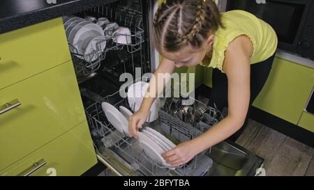 Schlaue Mädchen lernen, Geschirrspüler zu benutzen. Stilvolle, moderne Einbauküche in grünem Schwarz. Kind legt sauberes Geschirr. Stockfoto