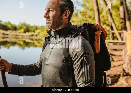 Mittlerer Erwachsener Mann mit Rucksack am See mit einem Stock. Männlicher Camper Camping am See im Wald. Stockfoto