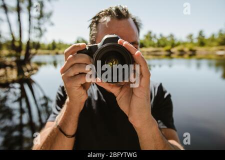 Reife Mann, der mit einer dslr-Kamera fotografiert. Männlicher Fotograf steht am See und fotografiert mit der Digitalkamera. Stockfoto