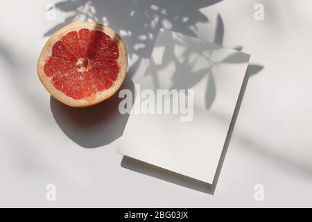 Sommer Schreibwaren Stillleben Szene. Nahaufnahme von geschnittenen Grapefruit-Früchten auf weißem Tischhintergrund bei Sonnenlicht. Leere Papierkarten, Einladungen Mockup Szene Stockfoto