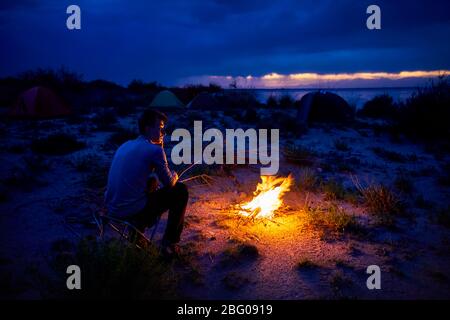 Mann, bei Lagerfeuer und Zelt in der Nacht am Strand des Sees Stockfoto