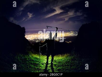 Mann mit Trekking Pole an der Spitze am Nachthimmel mit Sternen und leuchtende Stadt Licht unter Stockfoto