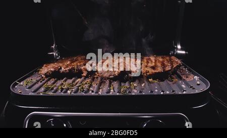 Köstliches, saftiges Steak-Kochen auf dem Grill. Braten Sie das Rindfleisch auf einem elektrischen Bräter, Rosmarin, schwarzem Pfeffer und Salz. Zeitlupe. Stockfoto