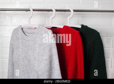 Die Kleidung hängt an Kleiderbügeln. Pullover in Rot, Grün und Melange. Mode und Stil, Schaufenster, Store, Konzept. Weiße Ziegelwand, Komfort und Sauberkeit Stockfoto