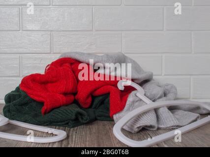 Ein Haufen Kleider gegen eine weiße Ziegelwand. Verstreute Kleiderbügel. Mode, Stil. Pullover in Rot, Grün und Melange. Chaos, Hausroutine Stockfoto