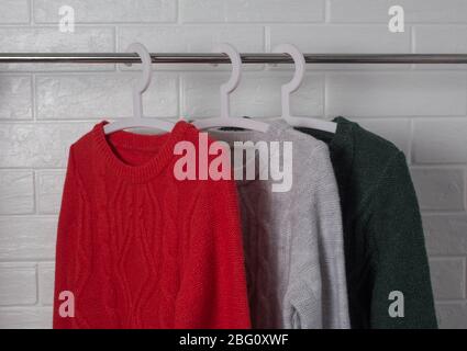 Die Kleidung hängt an Kleiderbügeln. Pullover in Rot, Grün und Melange. Mode und Stil, Schaufenster, Store, Konzept. Weiße Ziegelwand, Komfort und sauber Stockfoto
