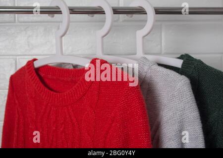 Die Kleidung hängt an Kleiderbügeln. Pullover in Rot, Grün und Melange. Mode und Stil, Schaufenster, Store, Konzept. Weiße Ziegelwand, Komfort Stockfoto