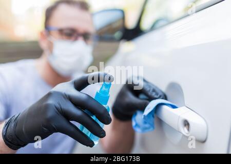 Fahrer mit Gesichtsmaske trägt Gummischutzhandschuhe wischen und sprühen Autotür Griff. Konzept zur Virus- und Bakterienprävention. Stockfoto