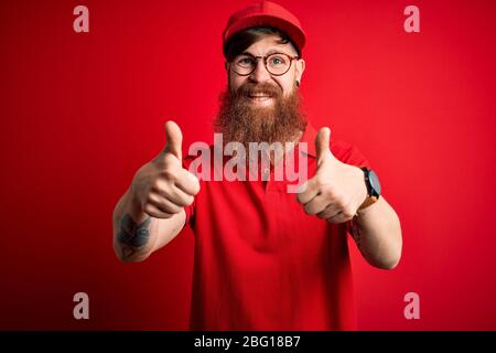 Junge schöne Lieferung Mann trägt Brille und rote Kappe über isolierten Hintergrund Erfolg Zeichen tun positive Geste mit der Hand, Daumen hoch lächelnd und Stockfoto