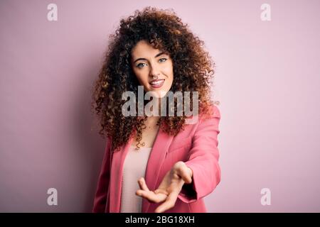 Junge schöne Geschäftsfrau mit lockigen Haaren und Piercing tragen elegante Jacke lächelnd fröhlich bietet Handfläche Hand geben Hilfe und Akzeptanz Stockfoto