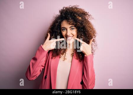 Junge schöne Geschäftsfrau mit lockigen Haaren und Piercing trägt elegante Jacke lächelnd fröhlich zeigen und zeigen mit den Fingern Zähne und Mund. Stockfoto