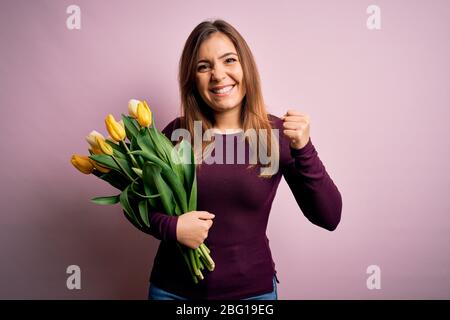 Junge blonde Frau mit romantischen Bouquet von gelben Tulpen Blumen über rosa Hintergrund sehr glücklich und aufgeregt tun Gewinner Geste mit erhobenen Armen Stockfoto