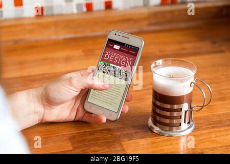 Responsive Design Webseite. Mädchen mit einer Kaffeetasse und einem Smartphone in der Hand. Auf dem Bildschirm des Mobiltelefons gibt es eine ansprechende Website von Kaffee Stockfoto