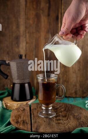 Milch wird in den Karaffe gegossen. Geyser Kaffeemaschine, Kaffeesahne auf einem Holztisch. Glasbecher mit Cappuccino zur Kaffeebereitung. Kaffee zubereiten, Hand Stockfoto