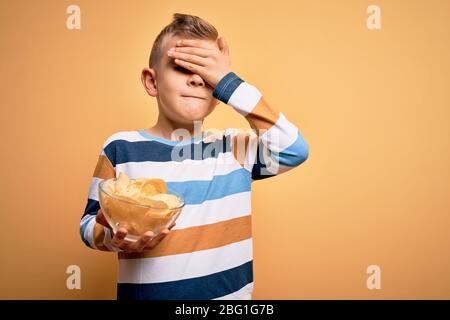 Junge kleine kaukasische Kind essen unheisse Kartoffeln Chips Chips über gelbem Hintergrund gestresst mit Hand auf Kopf, schockiert vor Scham und Überraschung f Stockfoto