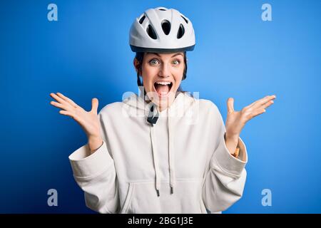 Junge schöne Rotschopf Radfahrer Frau trägt Fahrradhelm über isoliert blauen Hintergrund feiern verrückt und erstaunt für den Erfolg mit erhobenen Armen und Stockfoto