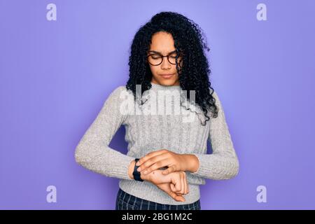 Junge afroamerikanische Frau in lässigen Pullover und Brille über lila Hintergrund Überprüfung der Zeit auf Armbanduhr, entspannt und zuversichtlich Stockfoto