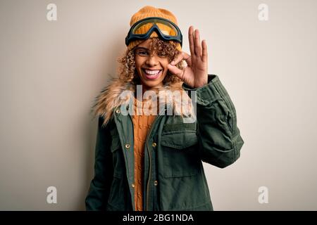 Junge afroamerikanische Skifahrer Frau mit lockigen Haaren tragen Schnee Sportbekleidung und Skibrille lächelnd positiv tun ok Zeichen mit Hand und Finger. Succe Stockfoto