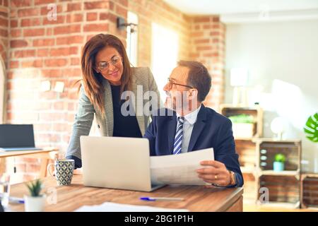 Zwei Geschäftsleute im mittleren Alter lächeln glücklich und zuversichtlich. Arbeiten Sie im Büro mit einem Lächeln im Gesicht mit einem Laptop zusammen Stockfoto