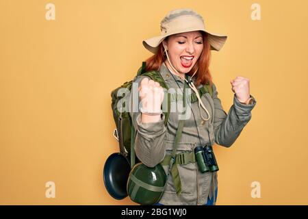Junge Rotschopf Backpacker Frau Wandern mit Rucksack und Hut über gelben Hintergrund feiern überrascht und erstaunt für den Erfolg mit Armen angehoben ein Stockfoto