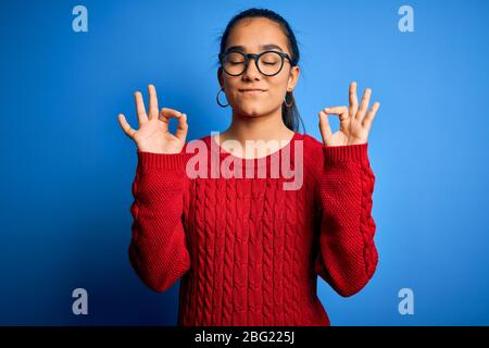 Junge schöne asiatische Frau mit lässigen Pullover und Gläser über blauem Hintergrund entspannen und lächeln mit geschlossenen Augen tun Meditation Geste mit f Stockfoto