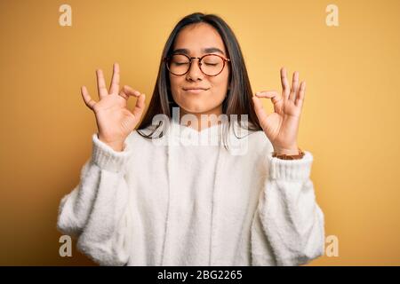 Junge schöne asiatische Frau mit lässigen Pullover und Gläser über gelbem Hintergrund entspannen und lächeln mit geschlossenen Augen tun Meditation Geste mit Stockfoto
