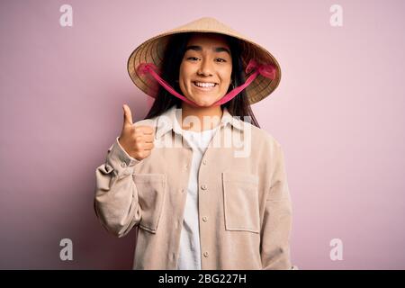 Junge schöne Frau trägt traditionelle konische asiatische Hut über isoliert rosa Hintergrund tun glücklich Daumen nach oben Geste mit der Hand. Genehmigung von Expressio Stockfoto