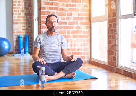 Mittelalter schöner Sportler sitzt auf Matte tun Stretching Yoga Übung im Fitnessstudio machen Fisch Gesicht mit Lippen, verrückt und komische Geste. Witziger Ausdruck Stockfoto