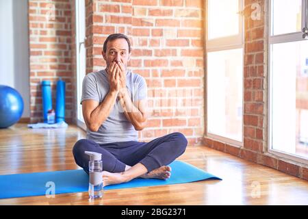 Mittelalter schöner Sportler sitzt auf Matte tun Stretching Yoga Übung im Fitnessstudio lachen und verlegen kichern Mund mit den Händen, Klatsch an Stockfoto