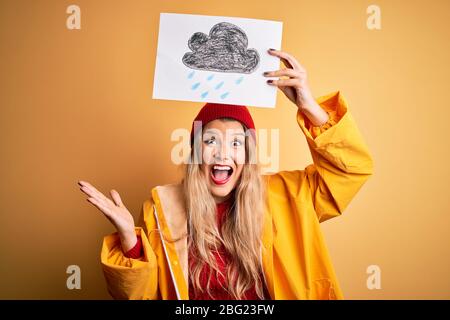 Junge schöne blonde Frau trägt Regenmantel und Wollmütze mit Banner mit Wolke Bild sehr glücklich und aufgeregt, Gewinner Ausdruck feiert victo Stockfoto