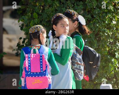 Drei junge turkmenische Mädchen tragen das grüne Nationalkleid, die von der Schule in Turkmenistan gehen. Gruppe turkmenischer Kinder, die mit wütender Gesichte zurückblicken Stockfoto