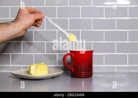Mans Hand hält einen Teelöffel Butter mit einem roten Kaffeebecher, um kugelsicheren Kaffee zu machen. Stockfoto
