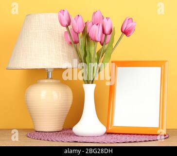 Bunte Bilderrahmen, Lampe und Blumen auf Holztisch auf gelbem Hintergrund Stockfoto