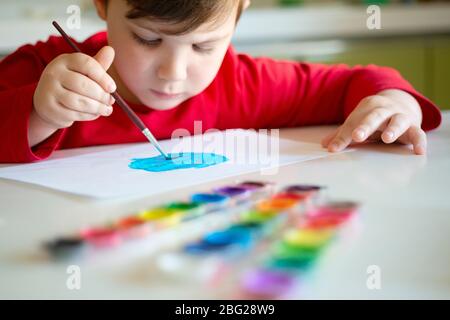 Der Junge lernt, mit einem Pinsel auf Papier zu malen, der an einem Tisch sitzt Stockfoto