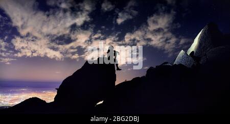 Mann mit Dreadlocks sitzen auf dem Felsen in Silhouette in der Nacht Himmel mit Sternen und leuchtende Stadt hellen Hintergrund. Stockfoto