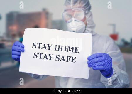 Arzt in einem Schutzanzug bei der medizinischen Masken tragen, halten Papier mit Text ZU HAUSE BLEIBEN SICHER. Zu Hause bleiben in Selbstisolation politische Kampagne zu cont Stockfoto