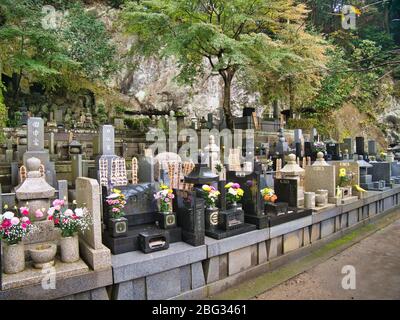 Vor dem Hintergrund eines Felsvorstieges und von Bäumen schmücken farbenfrohe Blumen die gut gehüteten Grabsteine auf einem Friedhof in der Nähe von Kamakura, nahe Tokio, Japan. Stockfoto